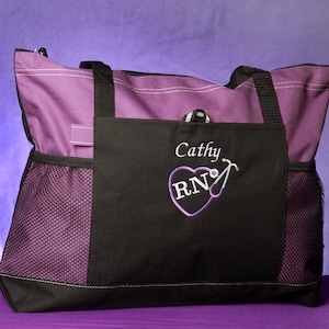 May include: Purple and black tote bag with white stitching and black handles. The front pocket features the name "Cathy" and the letters "RN" inside a heart with a stethoscope design. Mesh side pockets.