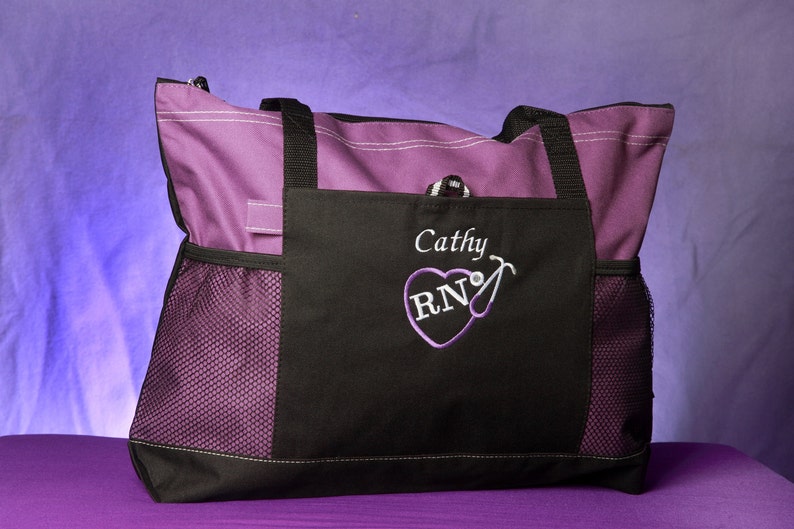 Peut inclure: Sac fourre-tout violet et noir avec une poche en filet et un c&oelig;ur brod&eacute; blanc avec le texte "RN" et le nom "Cathy" au-dessus.