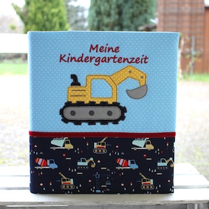 Könnte beinhalten: Eine blaue Aufbewahrungsbox mit weißen Punkten und einer gelben Bagger-Applikation. Der Text "Meine Kindergartenzeit" ist rot gestickt. Der untere Teil zeigt ein Muster von Marineblau-Baufahrzeugen.