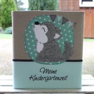 Könnte beinhalten: Eine rechteckige Aufbewahrungsbox mit grauem, leinenartigem Äußeren. Die Box hat unten ein türkis-weißes Vichy-Muster und eine runde Applikation mit einem heulenden Wolf und Sternenmuster. Der Text "Meine Kindergartenzeit" wird angezeigt.
