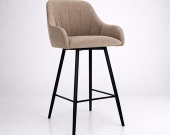 May include: A bar stool featuring a beige upholstered seat with vertical channel tufting and integrated armrests. The seat is supported by four slender, matte black metal legs connected by a square footrest frame, providing a stable base.