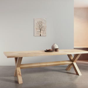 May include: A light wood dining table with a cross-shaped base and a shelf. The table is in a modern living room with a light-colored wall and a painting of a tree on the wall.