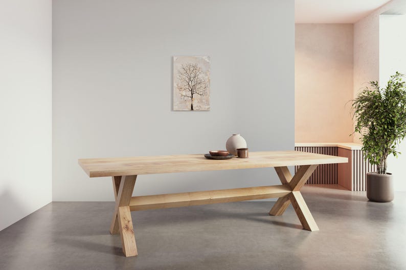 May include: A light wood dining table with a cross-shaped base and a shelf. The table is in a modern living room with a light-colored wall and a painting of a tree on the wall.