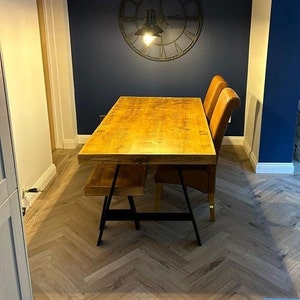 May include: A wooden dining table with a natural finish and black metal trestle legs. The table is set with two brown leather chairs and a matching bench. The table is in a room with a dark blue wall and a large clock on the wall.