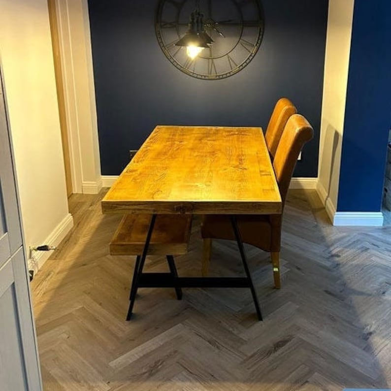May include: A wooden dining table with a natural finish and black metal trestle legs. The table is set with two brown leather chairs and a matching bench. The table is in a room with a dark blue wall and a large clock on the wall.