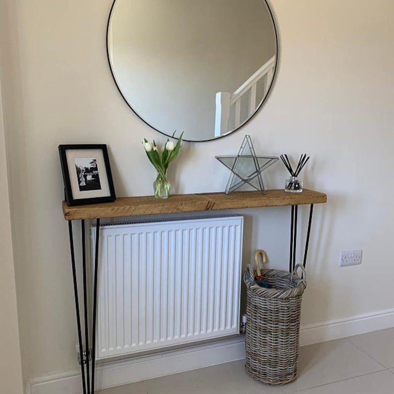 K&ouml;nnte beinhalten: Ein rustikaler Holz-Konsolentisch mit schwarzen Haarnadelbeinen. Der Tisch ist mit einem gerahmten Foto, einer Vase mit wei&szlig;en Blumen, einem Glasstern und einem Diffusor dekoriert. Hinter dem Tisch befindet sich ein wei&szlig;er Heizk&ouml;rper, und auf dem Boden steht ein Weidenkorb.