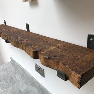 May include: A rustic wooden shelf with a live edge and a natural wood finish. The shelf is supported by four black metal brackets. The shelf is mounted on a white wall.