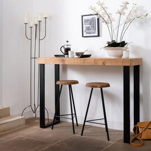 May include: A wooden console table with black metal legs and a wood top. Two black metal stools are placed in front of the table. A white vase with white orchids is on the table. A black and white framed picture is on the wall behind the table.