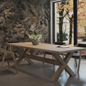 May include: A wooden dining table with a natural finish and a cross-shaped base. The table is surrounded by four chairs with black upholstery. The chairs have a similar wooden frame with a natural finish. The table is set in a room with a stone wall and a large window overlooking a fall foliage scene.