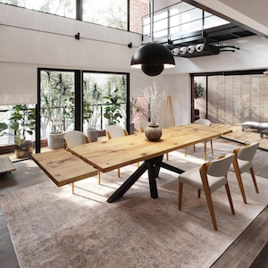 May include: A large, light-coloured wooden dining table with a black base and two extension leaves. The table is surrounded by six chairs with light-coloured seats and wooden legs. The room has large windows, a rug, and a black pendant light.