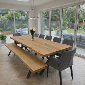 Könnte beinhalten: Ein großer Esstisch aus hellem Holz mit einer Baumkante, dazu eine passende Bank und mehrere grau gepolsterte Stühle. Der Tisch ist etwa 305 cm lang. Der Raum hat große Fenster mit Blick auf einen Garten.