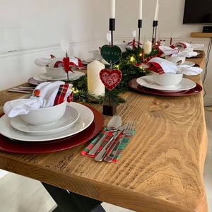 May include: A rustic wooden table with a black X-shaped base is set for a holiday dinner. The table is decorated with a garland of greenery, red and white plaid napkins, white bowls, and red chargers. There are candlesticks with white candles and heart-shaped decorations with the text "For You".