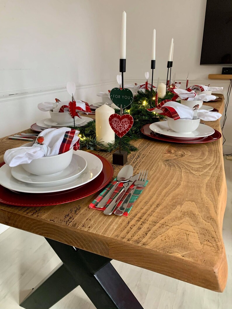 May include: A rustic wooden table with a black X-shaped base is set for a holiday dinner. The table is decorated with a garland of greenery, red and white plaid napkins, white bowls, and red chargers. There are candlesticks with white candles and heart-shaped decorations with the text "For You".
