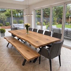 May include: A large rectangular dining table with a natural live edge wood top and black metal legs, accompanied by a matching long wooden bench and several dark grey upholstered chairs with black legs, featuring a glass vase of white roses.