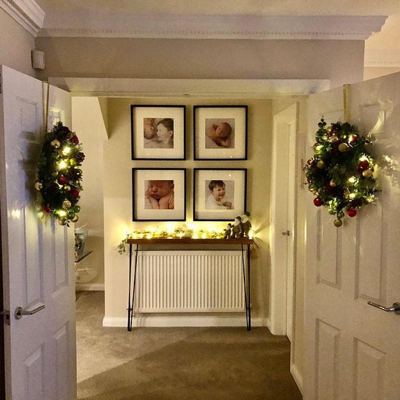 K&ouml;nnte beinhalten: Ein Flur mit einem wei&szlig;en Heizk&ouml;rper, einem Holz-Konsolentisch mit einem schwarzen Metallrahmen und vier schwarzen gerahmten Fotos an der Wand. Die Fotos zeigen Babys und Kinder. An den T&uuml;ren h&auml;ngen zwei Weihnachtskr&auml;nze.