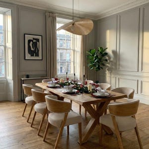 May include: A dining room scene featuring a large wooden dining table set for a meal. Eight beige upholstered chairs surround the table. An abstract artwork hangs on the wall, and a unique light fixture hangs above.