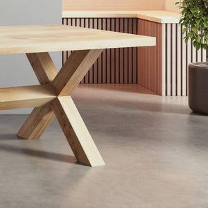May include: A light wood table with a cross-shaped base. The table has a natural wood finish and is sitting on a light gray concrete floor.