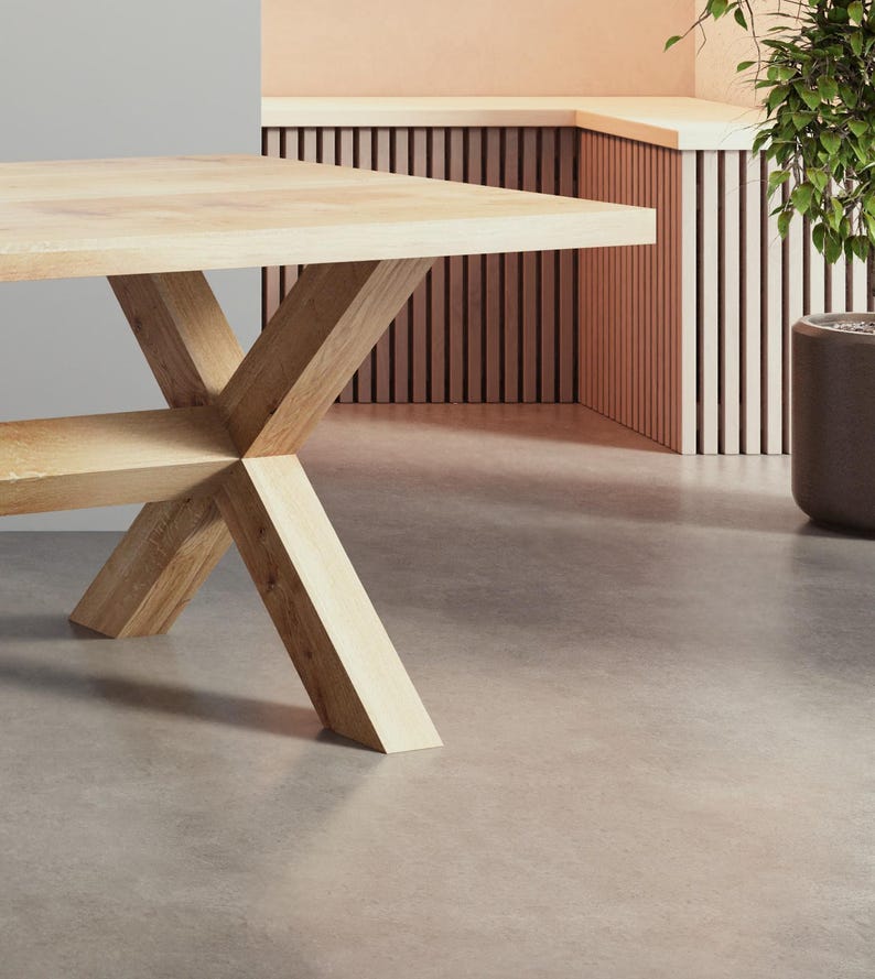 May include: A light wood table with a cross-shaped base. The table has a natural wood finish and is sitting on a light gray concrete floor.