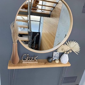 Op de afbeelding: Een ronde spiegel met houten frame hangt boven een houten plank. Op de plank staan een houten vaas met gedroogd pampasgras, een metalen "Glück" bord en een witte geometrische vaas met gedroogde palmbladeren. De muur is grijs geschilderd.