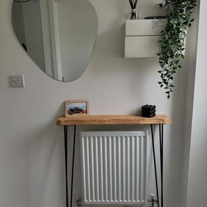 Op de afbeelding: Een houten consoletafel met zwarte haarspeldpoten. De tafel staat voor een witte radiator en heeft een kleine ingelijste foto en een zwart decoratief object erop.