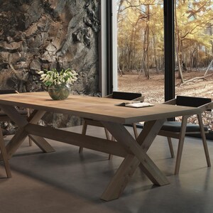 May include: A wooden dining table with a natural finish and a cross-shaped base. The table is set with four chairs, two of which are visible in the image. The chairs are upholstered in black fabric and have a simple design. The table is set against a backdrop of a stone wall and a window with a view of a forest.