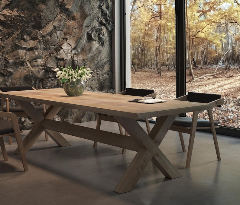 May include: A wooden dining table with a natural finish and a cross-shaped base. The table is set with four chairs, two of which are visible in the image. The chairs are upholstered in black fabric and have a simple design. The table is set against a backdrop of a stone wall and a window with a view of a forest.