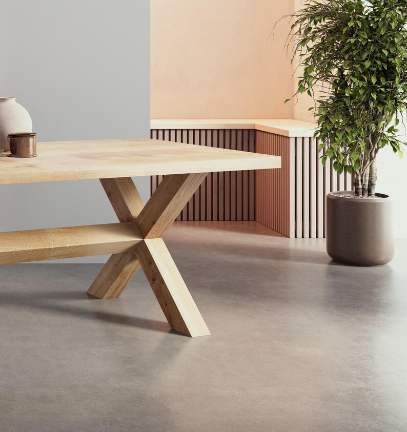 May include: A light wood dining table with a wide, rectangular top and an X-shaped base. The table is in a room with a light gray wall and a light peach wall with a wooden slatted bench.