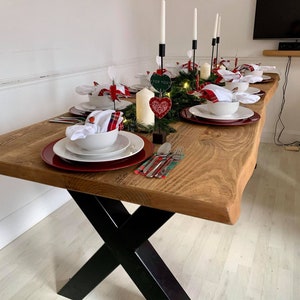 May include: A rustic wooden table with black X-shaped legs, set for a holiday dinner. The table is decorated with red and white plaid napkins, red and white heart ornaments, and white candles. The table is set with white plates, red chargers, and silverware.
