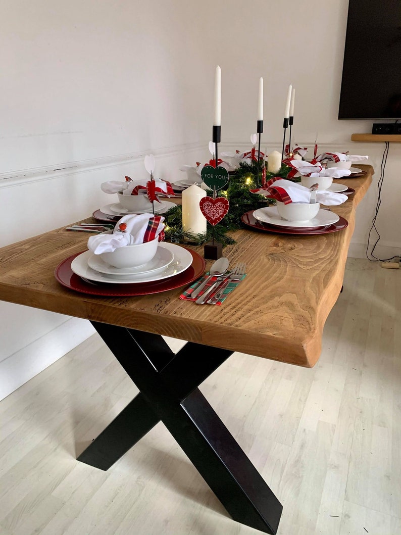 May include: A rustic wooden table with black X-shaped legs, set for a holiday dinner. The table is decorated with red and white plaid napkins, red and white heart ornaments, and white candles. The table is set with white plates, red chargers, and silverware.