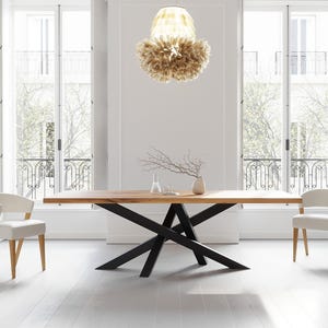 Op de afbeelding: Een moderne eetkamer met een grote houten tafel met zwarte metalen poten. Twee witte stoelen staan aan weerszijden. Boven de tafel hangt een decoratieve lamp. De kamer heeft grote ramen en een neutraal kleurenpalet.