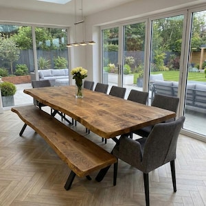 May include: A substantial wooden dining table with a live edge, accompanied by a bench and grey upholstered chairs. The table, approximately 300 cm long, rests on black metal legs. A vase of yellow flowers is in the center. Large windows surround the room.