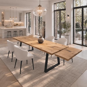 May include: A rectangular dining table with a natural wood grain top and black metal U-shaped legs. The table features an extendable leaf on one end and is surrounded by six white upholstered chairs with black legs, set in a bright open room.