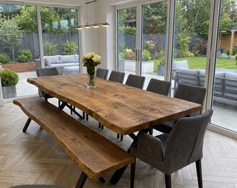 May include: A large rectangular dining table with a natural live edge wood grain finish in the colour Tosca, paired with a matching wooden bench and several dark grey upholstered chairs with black legs, set in a bright room.