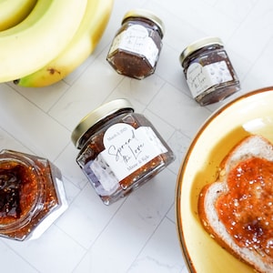 Op de afbeelding: Vier kleine glazen potjes met vijgenjam met witte etiketten. De etiketten hebben de tekst "Spread the Love" en "Mince de Figue". Een potje is open en een snee brood met vijgenjam zit in een gele kom.