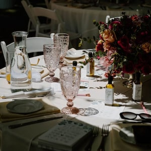 May include: A table setting with pink-tinted glassware, a floral arrangement, and small bottles of olive oil. The table is set for a special event, with plates, napkins, and silverware. Sunglasses and a menu are also visible.