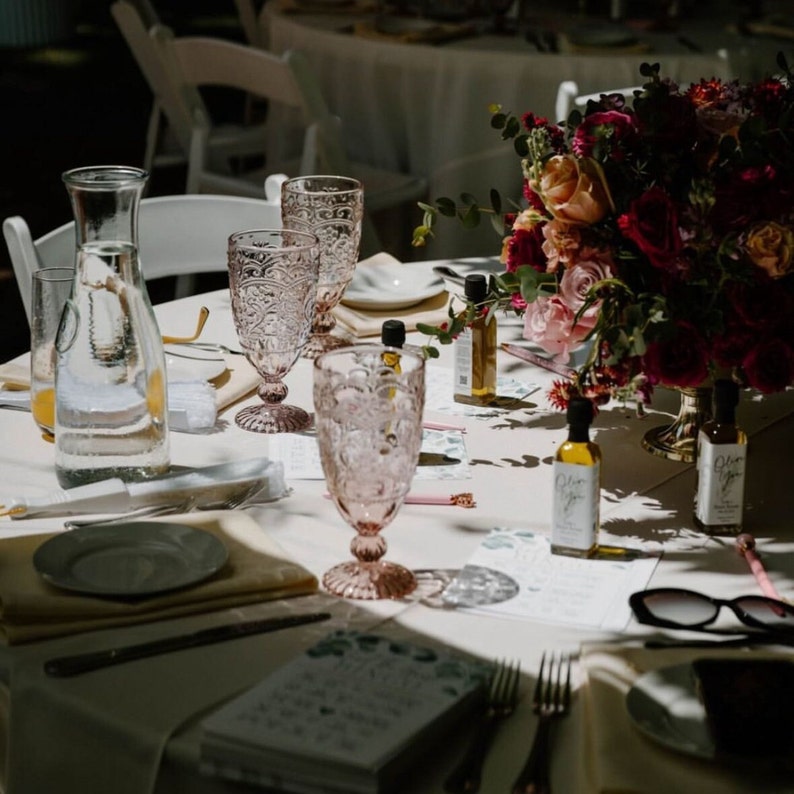 May include: A table setting with pink-tinted glassware, a floral arrangement, and small bottles of olive oil. The table is set for a special event, with plates, napkins, and silverware. Sunglasses and a menu are also visible.