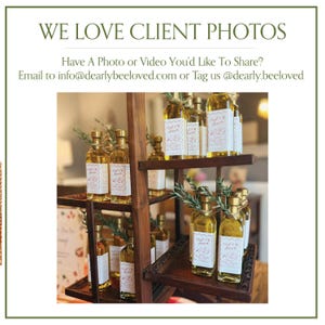 May include: A wooden display shelf holds several glass bottles filled with a golden liquid. Each bottle has a gold cap and a white label with pink floral accents. The text "WE LOVE CLIENT PHOTOS" is at the top.
