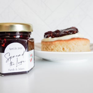 Op de afbeelding: Een klein glazen potje met zelfgemaakte jam met een wit etiket waarop "Spread the Love" staat met de datum "April 20, 2023" en de namen "Sarah & Adrian".