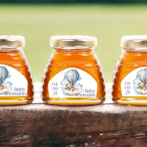 May include: Three small glass jars filled with honey, each with a white label featuring a cartoon bear in a blue hot air balloon. The labels also include the text "Baby Thomann" and the date "04 09 25".