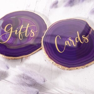 May include: Two purple agate coasters with gold trim. The coasters have the words "gifts" and "cards" written in gold lettering.