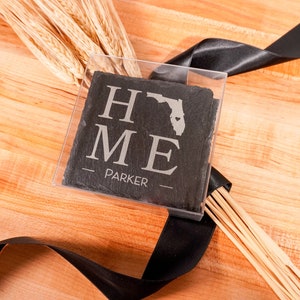 May include: A set of black slate coasters with a white inscription that reads "HOME" with a stylized outline of the state of Florida and a heart in the center. The text "PARKER" is below the state outline. The coasters are packaged in a clear plastic box with a black ribbon.
