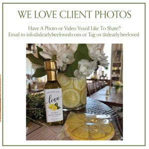 May include: A bottle of olive oil with a gold cap and a label that reads "Infused with love." The label also includes the text "Kirsten's Bridal Shower." A vase of flowers and a lemon-shaped cookie are also visible.