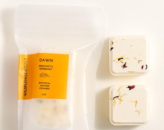 May include: A clear plastic bag with a yellow label reading "DAWN" and "Bergamot & Peppermint" with botanical shower steamers. Two square botanical shower steamers with colorful flower petals are next to the bag.