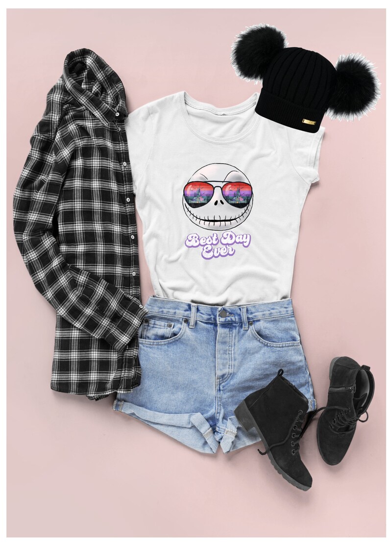 K&ouml;nnte beinhalten: Wei&szlig;es T-Shirt mit einem Grafikdruck von Jack Skellington, der eine Sonnenbrille mit einem Stadtbild-Reflex tr&auml;gt. Der Text "Best Day Ever" ist in Lila unter dem Grafikdruck gedruckt. Das T-Shirt wird mit Jeans-Shorts, einem schwarz-wei&szlig; karierten Hemd und schwarzen Stiefeln kombiniert.