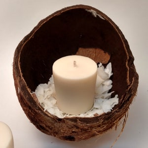 Puede incluir: Una vela blanca se encuentra en una cáscara de coco ahuecada, rodeada de copos de coco blancos. La cáscara de coco es marrón y tiene una textura rugosa.