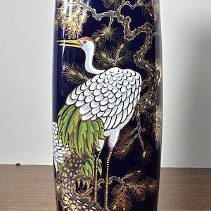 Könnte beinhalten: Eine hohe, marineblaue Keramikvase mit einer detaillierten Illustration eines Kranichs mit weißen Federn, grünen Akzenten und einem roten Kopf. Der Kranich ist vor einem Hintergrund aus goldfarbenen Kiefernzweigen und Blättern dargestellt.