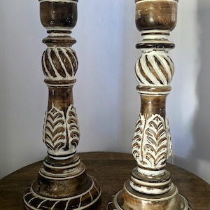 May include: Two tall wooden candle holders featuring a dark brown stained finish with white paint accents in the carved details. Each holder has a wide circular base, a central twisted sphere section, and a leaf-patterned column design.