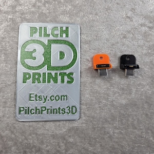 Può includere: L'immagine presenta due protezioni per porte USB-C, una arancione e una nera, accanto a un'etichetta argentata. L'etichetta riporta la scritta "PILCH 3D PRINTS" in verde, con "Etsy.com" e "PilchPrints3D" sotto. Lo sfondo è un tessuto grigio strutturato.