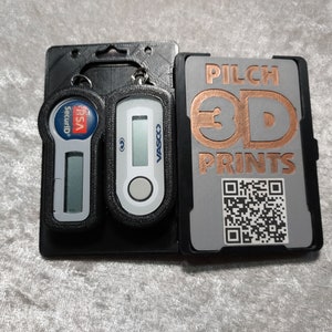 Può includere: Supporti neri stampati in 3D con due token di sicurezza e una carta. I token hanno display digitali e branding. La carta presenta il testo "PILCH 3D PRINTS" e un codice QR. Gli oggetti sono su una superficie grigio chiaro.