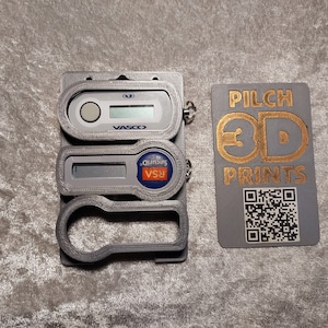 Può includere: Un porta token di sicurezza stampato in 3D di colore grigio. Il porta token ha un portachiavi e uno slot per una carta. La carta è grigia con un logo dorato che dice "PILCH 3D PRINTS" e un codice QR.